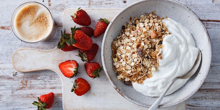 Yoghurt and muesli in a bowl with chopped strawberries and a hot latte on a wooden board.